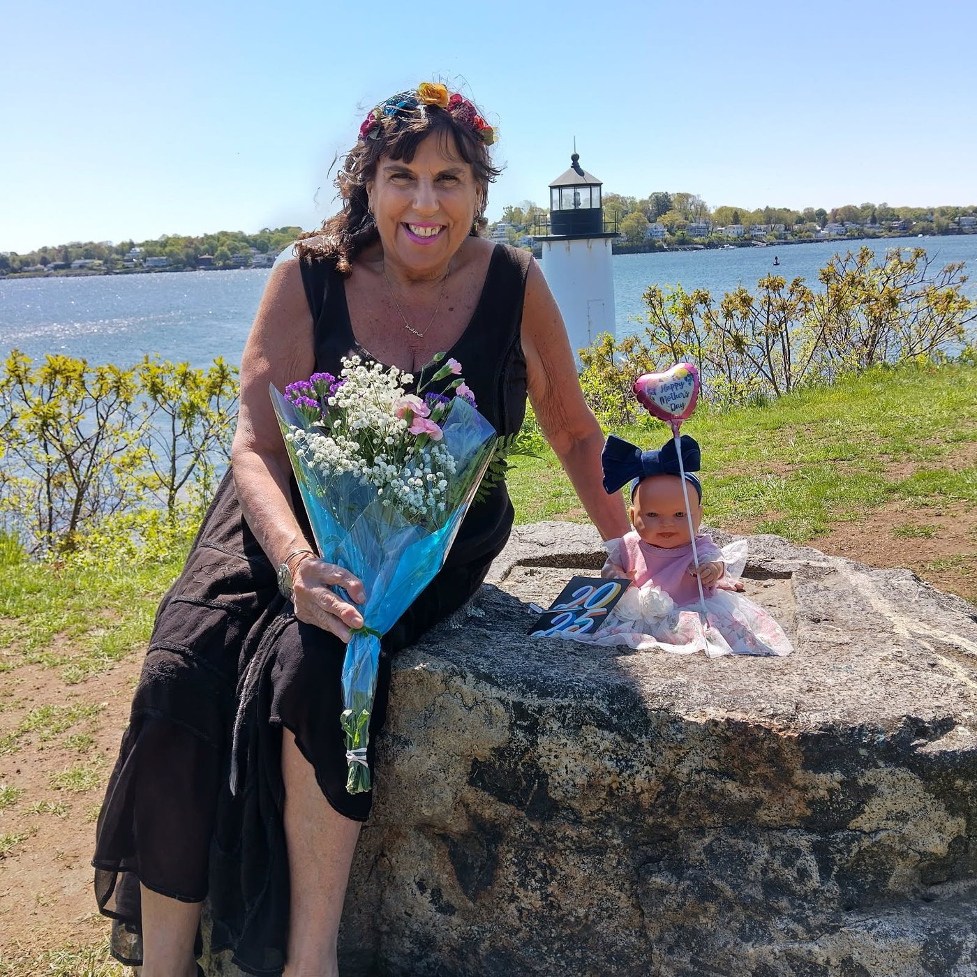 Rosemarie smiling by the water with a lighthouse behind her, holding flowers, with one of her dolls beside her